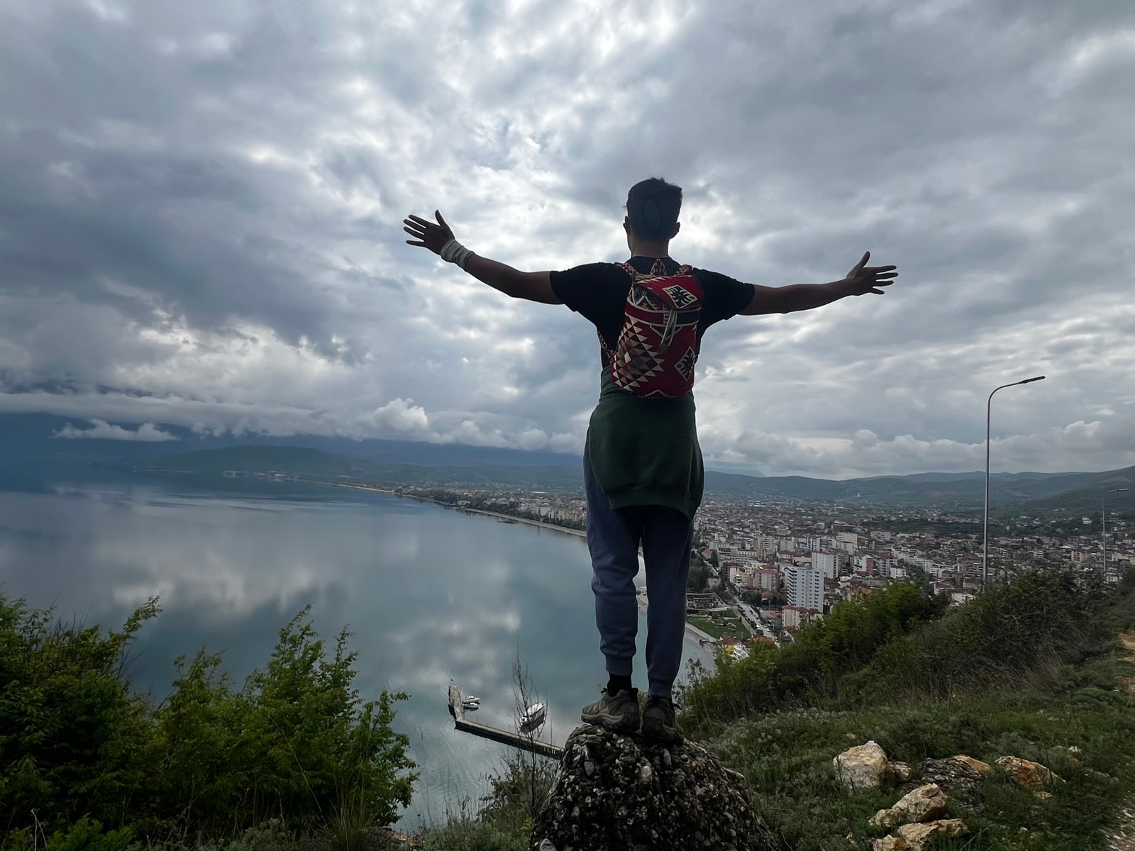 Hiker standing on a rock with arms spread wide, overlooking the city of Pogradec and Lake Ohrid from above